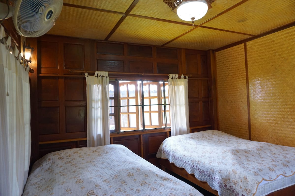 Ein Zimmer mit zwei Betten, Holzwänden, einem Deckenventilator und einem mit Vorhängen versehenen Fenster.
