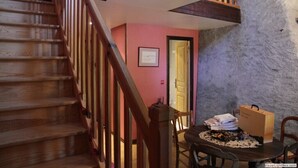 A staircase with wooden railings leading to a room with a pink wall and a stone wall. A table with a doily and a cardboard box is visible.