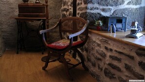 A rustic room with a stone wall, a wooden chair with a red cushion, a small table with a brass vase, and a blue heater.