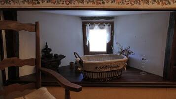 Une pièce chaleureuse avec une chaise en bois, une petite table sur laquelle se trouve un panier étiqueté « CAMPAGNOLO », une fenêtre ornée d’un rideau à motifs floraux et un vase rempli de fleurs.