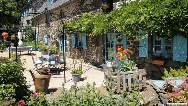 A stone building with blue shutters, a garden with potted plants, and outdoor seating.