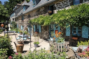 A stone building with blue shutters, a garden with potted plants, and outdoor seating.