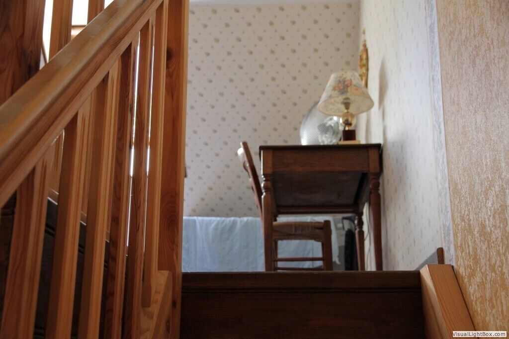 Una scala in legno conduce a una stanza con carta da parati decorata, una sedia di legno e un tavolo con una lampada.