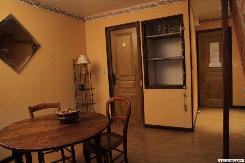 Uma área de refeições com mesa e cadeiras de madeira, uma tigela de comida, um abajur e uma porta com uma prateleira acima.