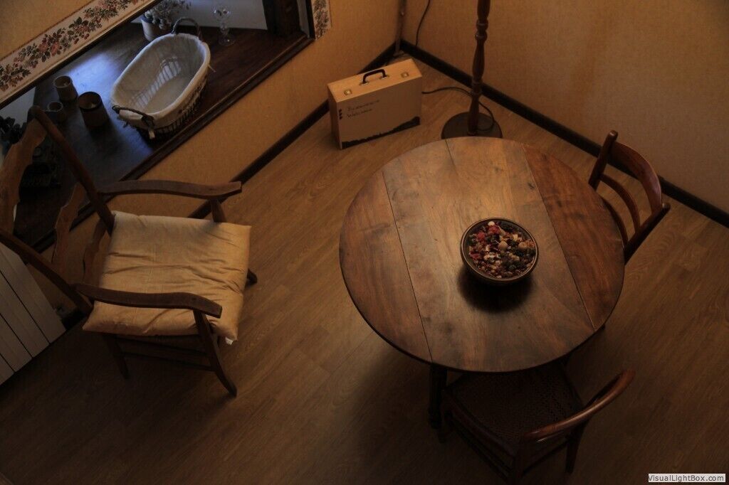 Une table à manger en bois, sur laquelle se trouve un saladier rempli de fruits, est entourée de chaises en bois. Un banc en bois est placé à l’extrémité de la table. Une petite boîte en bois se trouve sur le sol, à côté de la table.