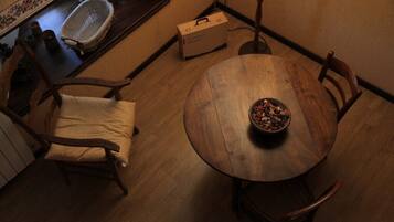 Une table à manger en bois, sur laquelle se trouve un saladier rempli de fruits, est entourée de chaises en bois. Un banc en bois est placé à l’extrémité de la table. Une petite boîte en bois se trouve sur le sol, à côté de la table.
