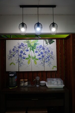 A room with a wooden wall, a green panel, a painting of blue flowers, a coffee maker, a glass pitcher, and a tray with cups. - Smile Mansion (Nakhon Si Thammarat)