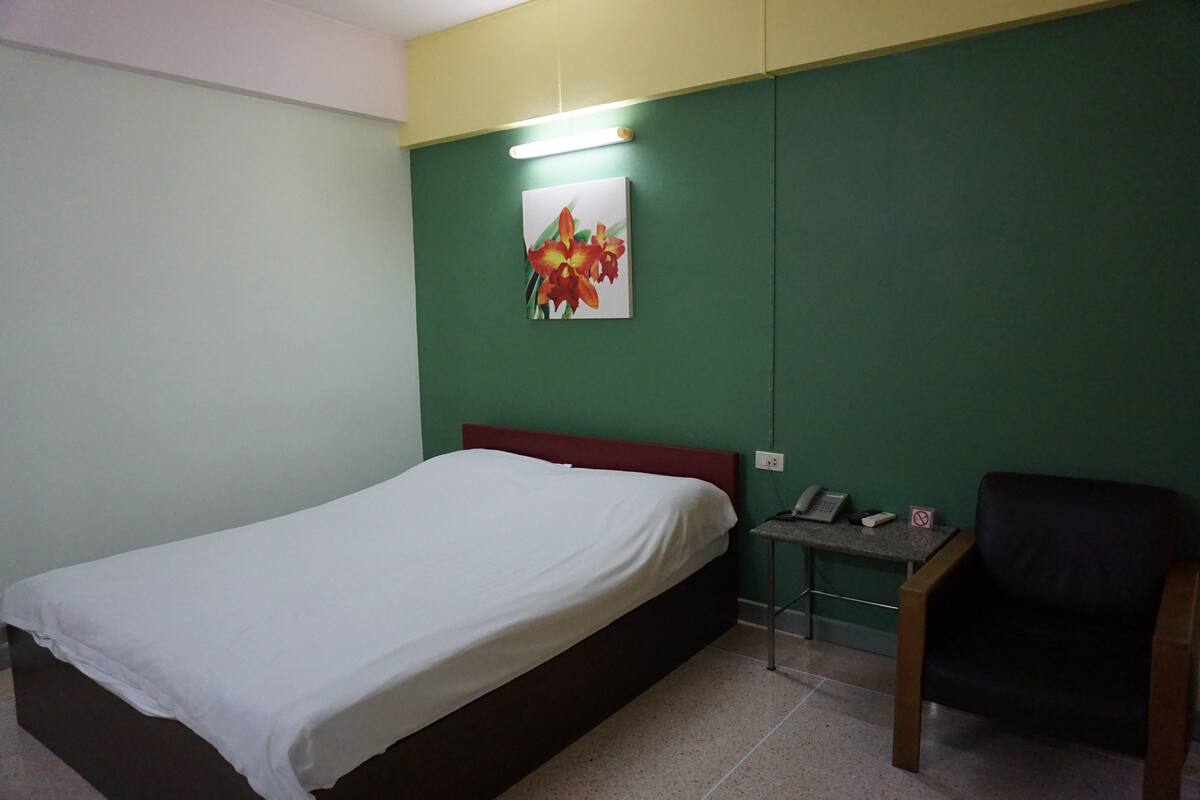 Un lit simple avec un drap blanc dans une pièce aux murs verts et à la tête de lit rouge. On y trouve une petite table avec un téléphone et un panneau, ainsi qu’une chaise noire sur la droite.