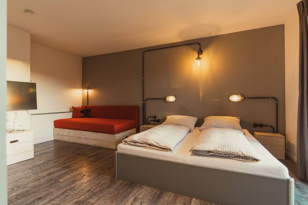 A bedroom with a bed, a red sofa, a nightstand, and a wall-mounted TV.