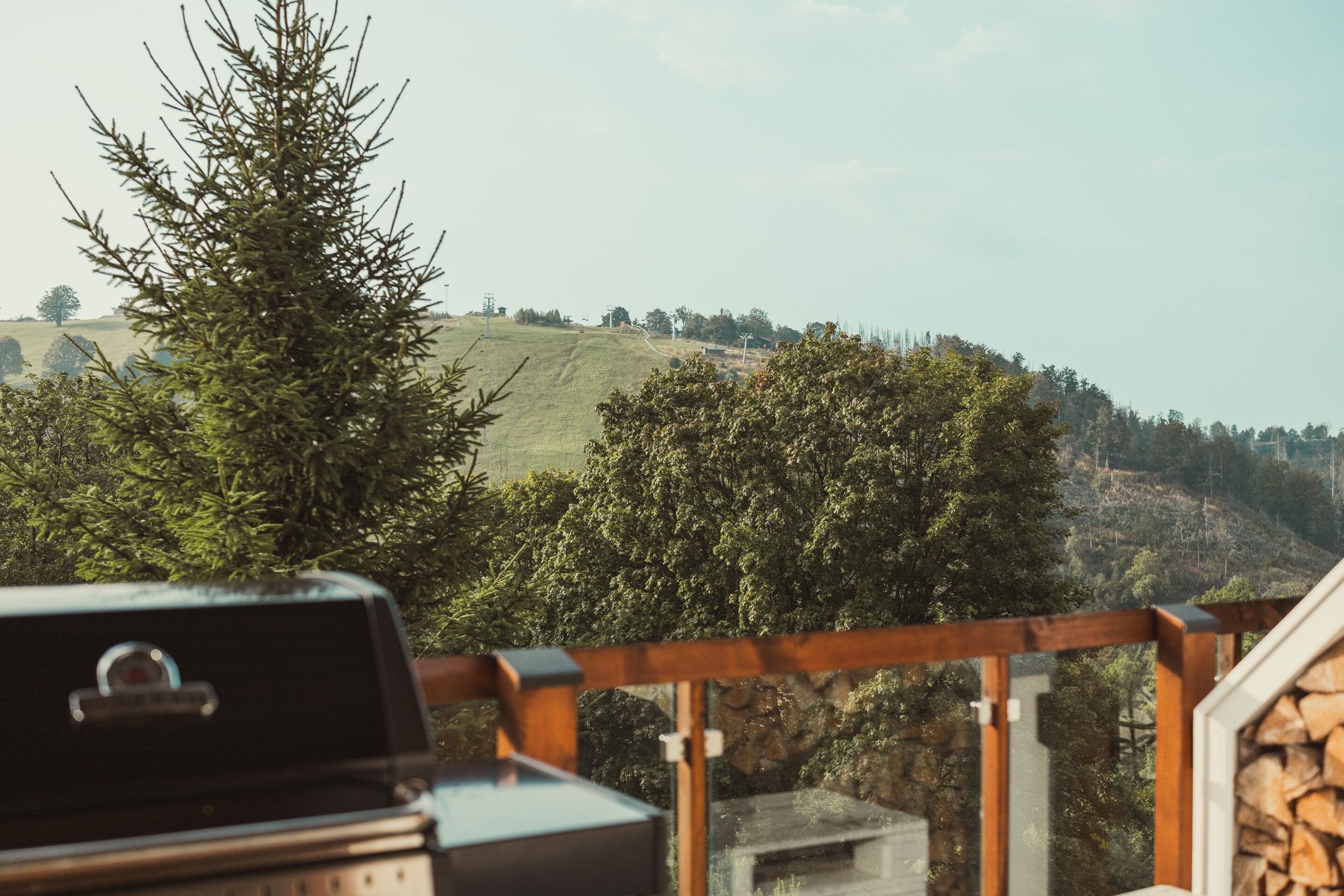 A balcony with a grill, overlooking a hilly landscape with dense trees and a clear sky.
