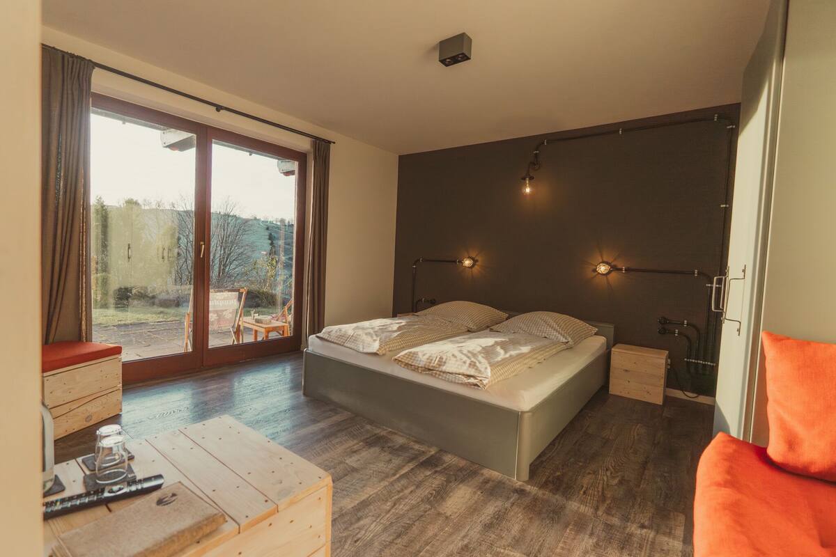 A bedroom with a large bed, a wooden nightstand, a small table, and a view through a sliding door to an outdoor area.