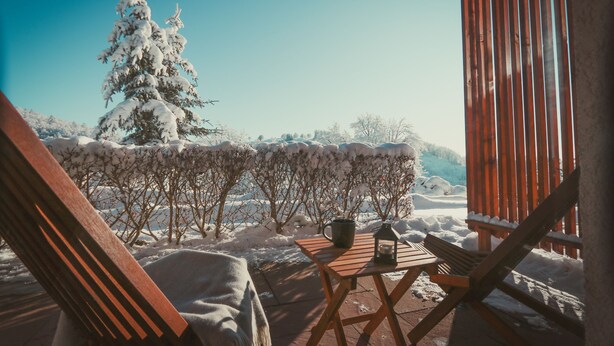 Una mesa de madera con un farol y una taza en un patio con nieve en el suelo y árboles al fondo.