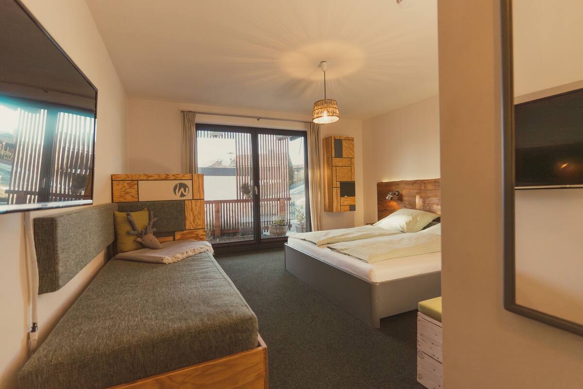 A compact hotel room with a bed, a sofa, a small table, and a window with blinds.