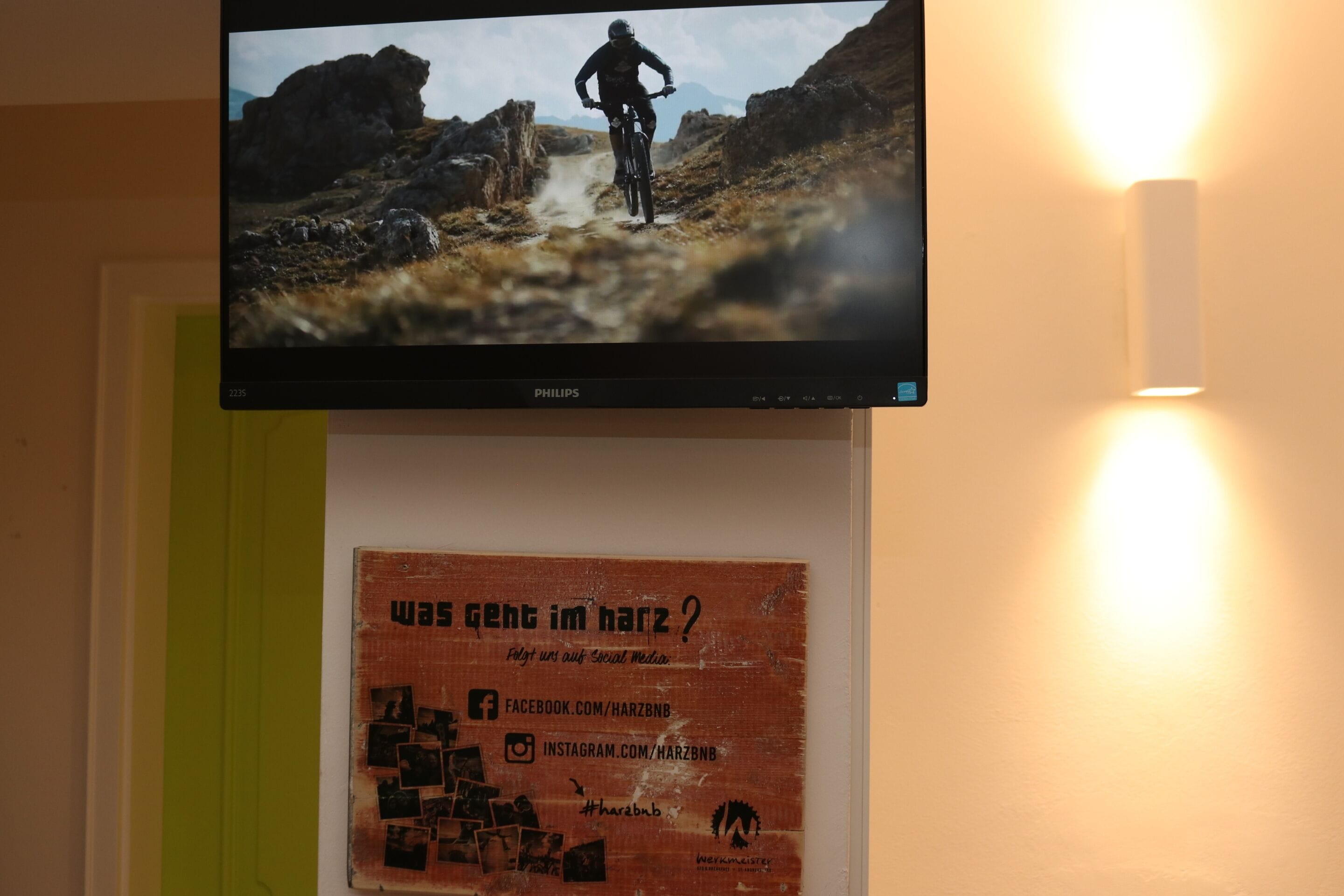 A television mounted on a wall displaying a mountain biking scene. Below the TV, there is a poster with text and social media icons.