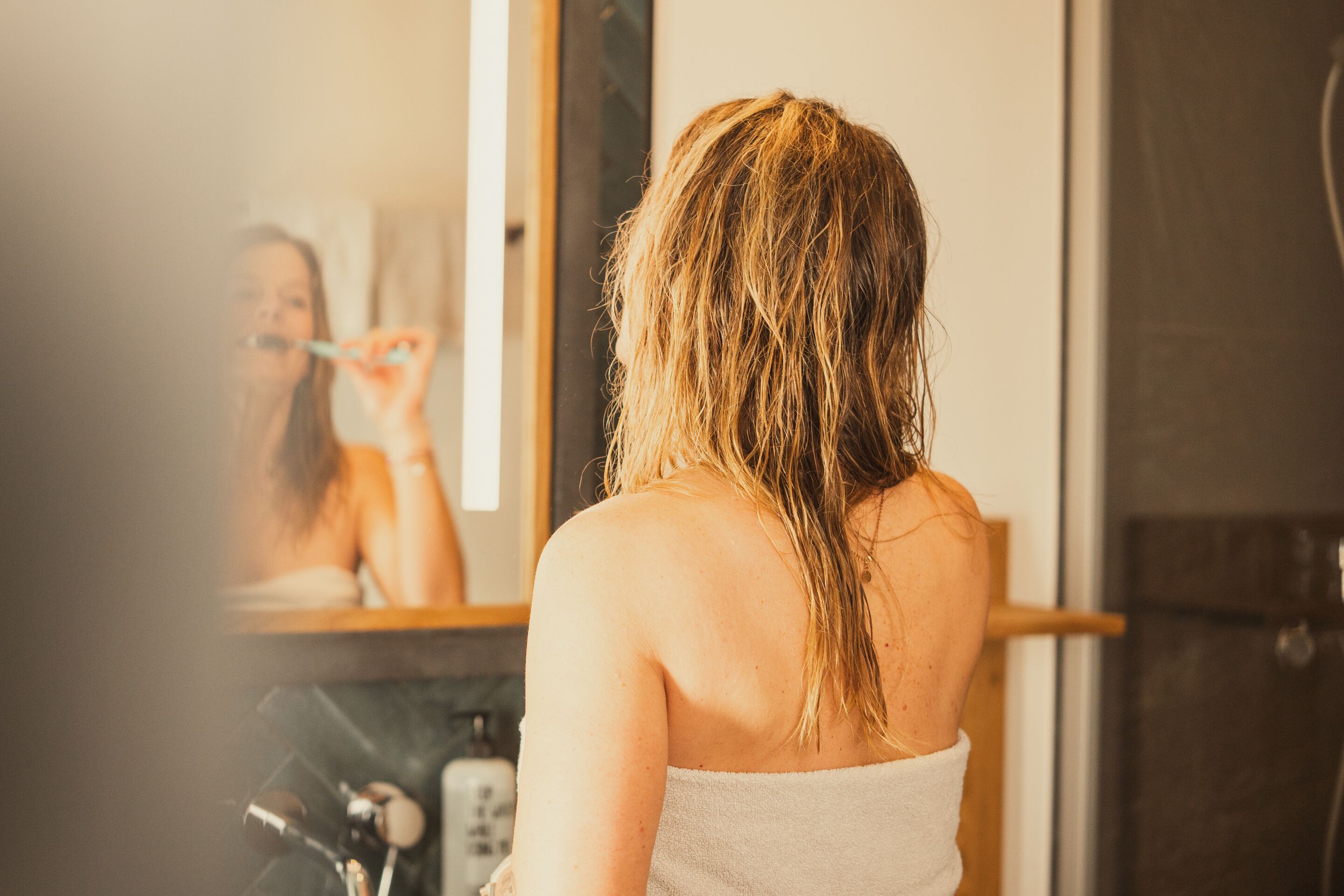 Une personne aux cheveux mouillés, dans une salle de bain, se regarde dans un miroir.