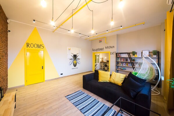 A modern interior with a yellow door labeled "ROOMS," a black sofa with yellow and green cushions, a hanging chair, and a bookshelf with books and plants.