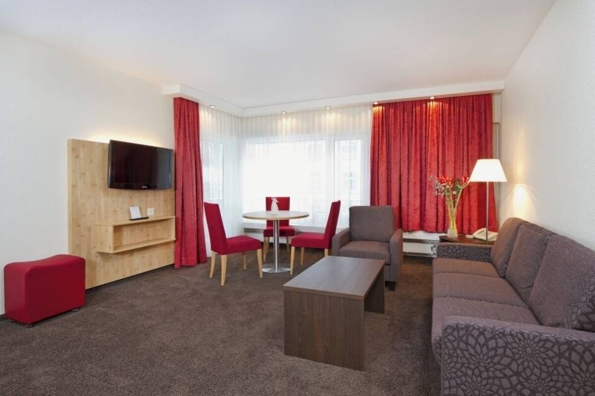 Una habitación de hotel moderna con un mueble de televisión de madera, un reposapiés rojo, un sofá gris, una mesa de comedor redonda con sillas rojas y una cortina roja.
