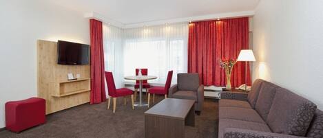 Una habitación de hotel moderna con un mueble de televisión de madera, un reposapiés rojo, un sofá gris, una mesa de comedor redonda con sillas rojas y una cortina roja.