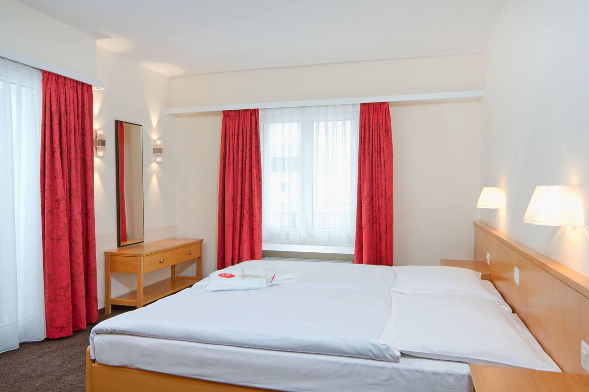 Una habitación de hotel con cama, mesita de noche de madera, espejo y cortinas rojas.