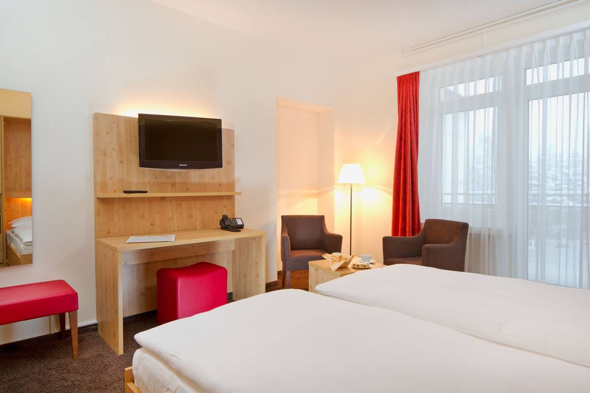 Habitación de hotel con cama, escritorio, sillas, televisor, lámpara y ventanas amplias.