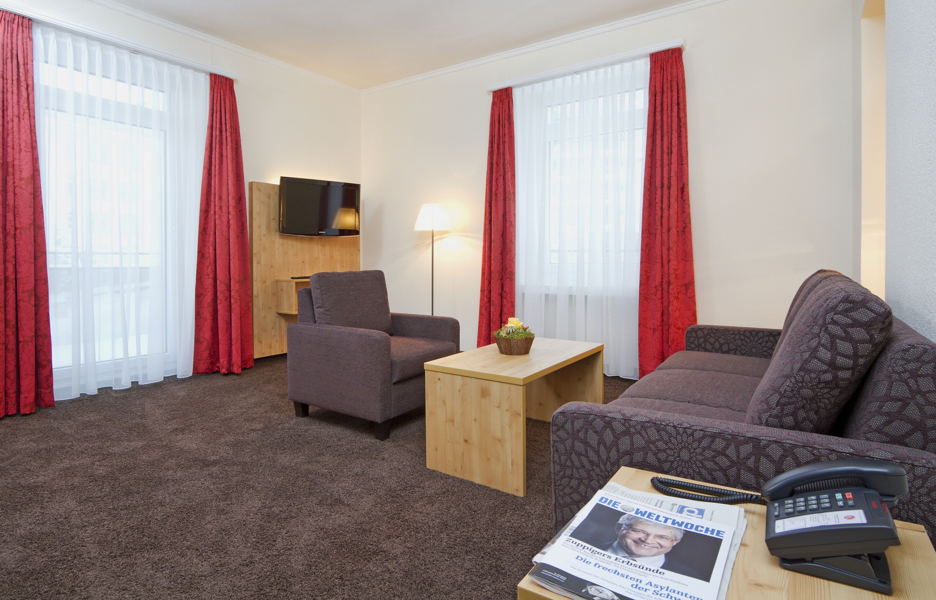 A hotel room with a sofa, armchair, wooden side table, lamp, and a flat-screen TV mounted on the wall.