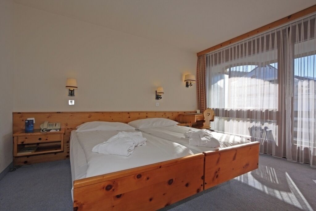 A hotel room with a wooden bed, two bedside tables, a nightstand, and a window with sheer curtains.