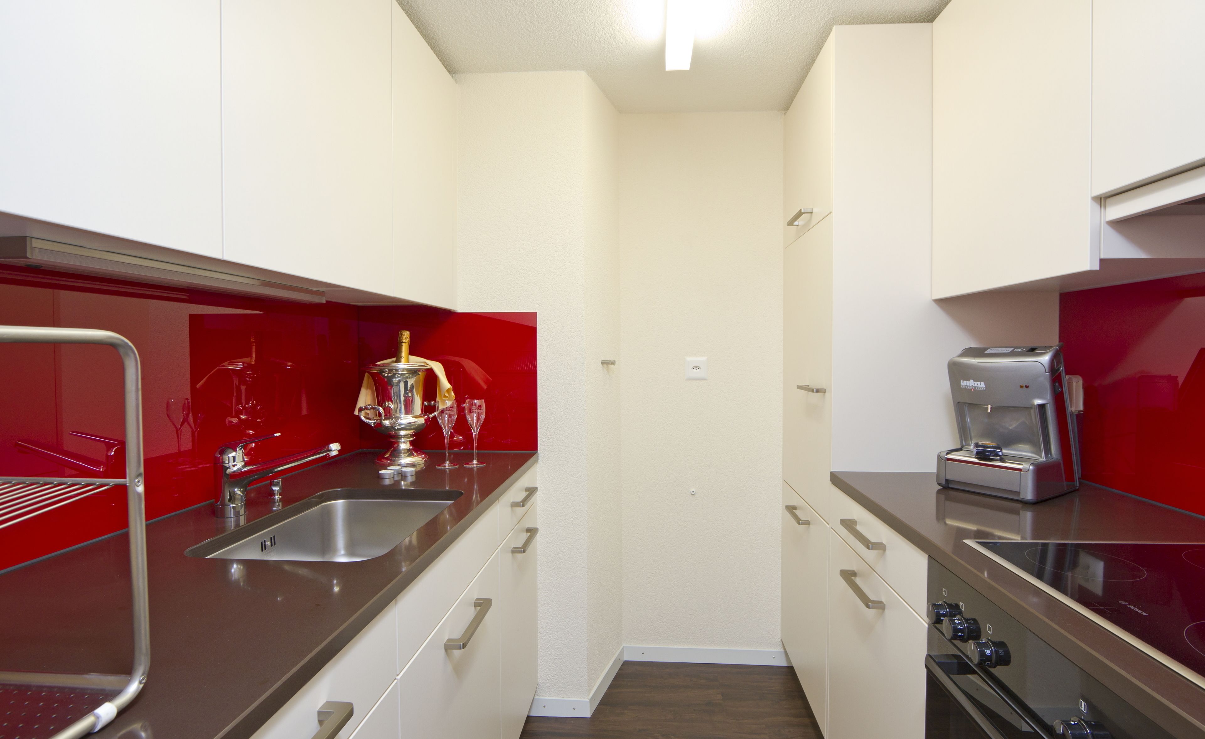 A compact kitchen with a stainless steel sink, a red backsplash, and a coffee machine.