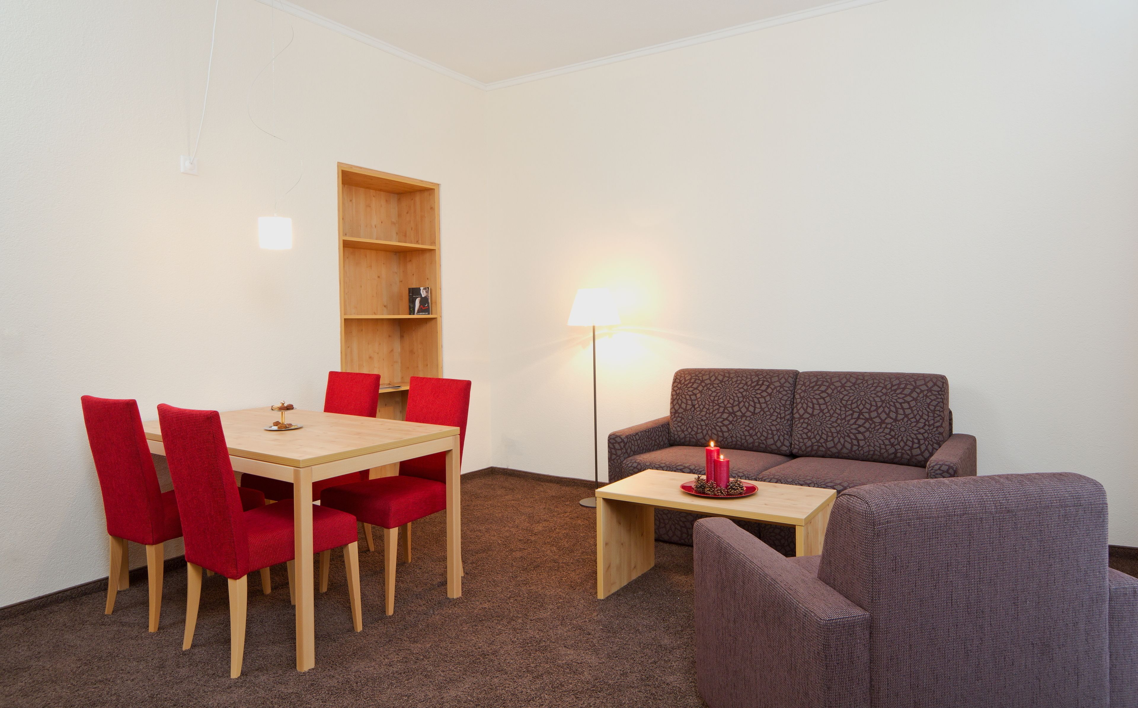 Une petite pièce meublée, avec une table et des chaises en bois, un canapé, une bibliothèque et un lampadaire.