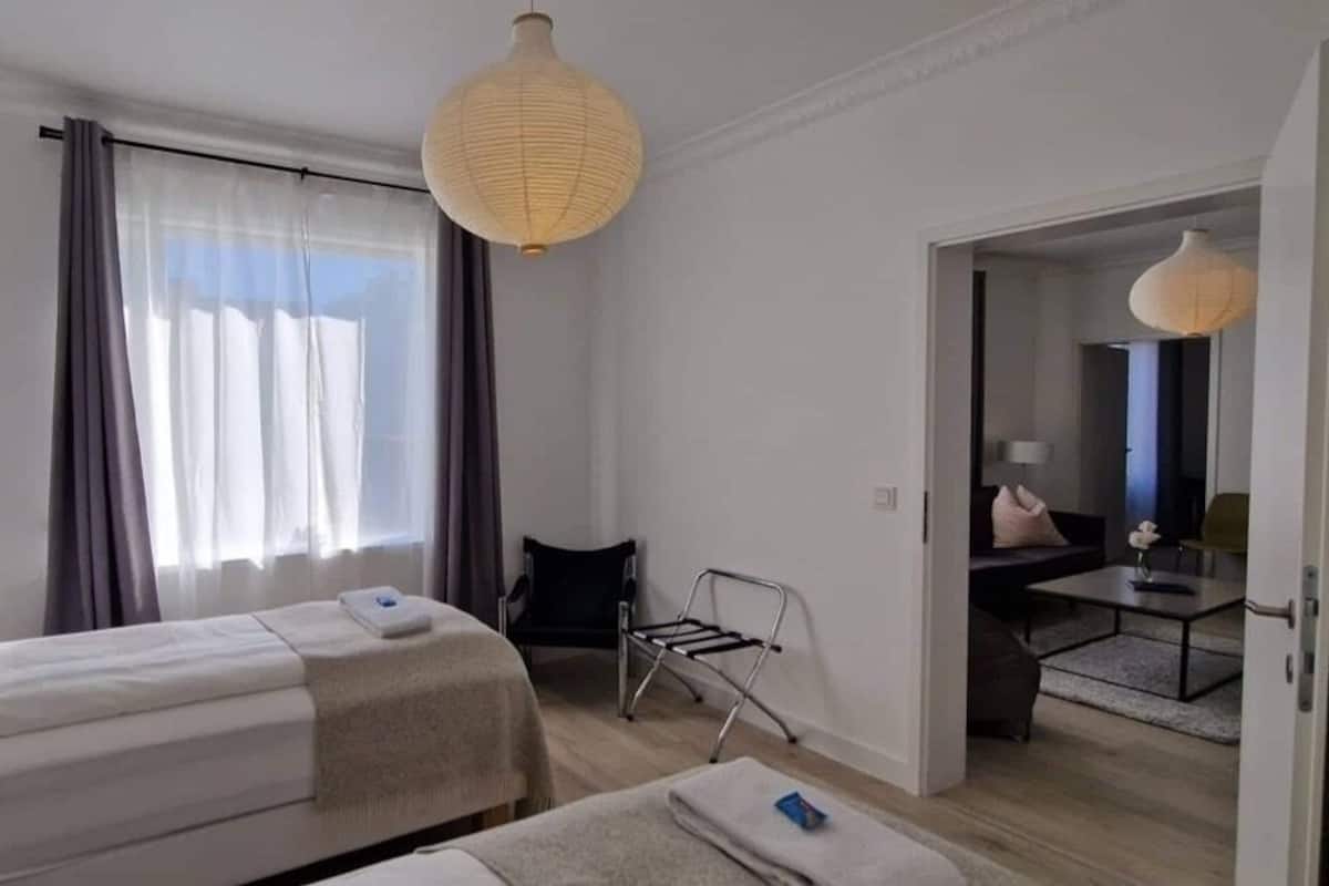 A hotel room with a bed, a chair, a table, and a lamp. There is a window with curtains and a door leading to another room.