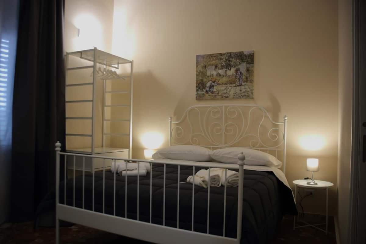 A bedroom with a bed, a bedside table with a lamp, a wall-mounted painting, and a wardrobe with a ladder.