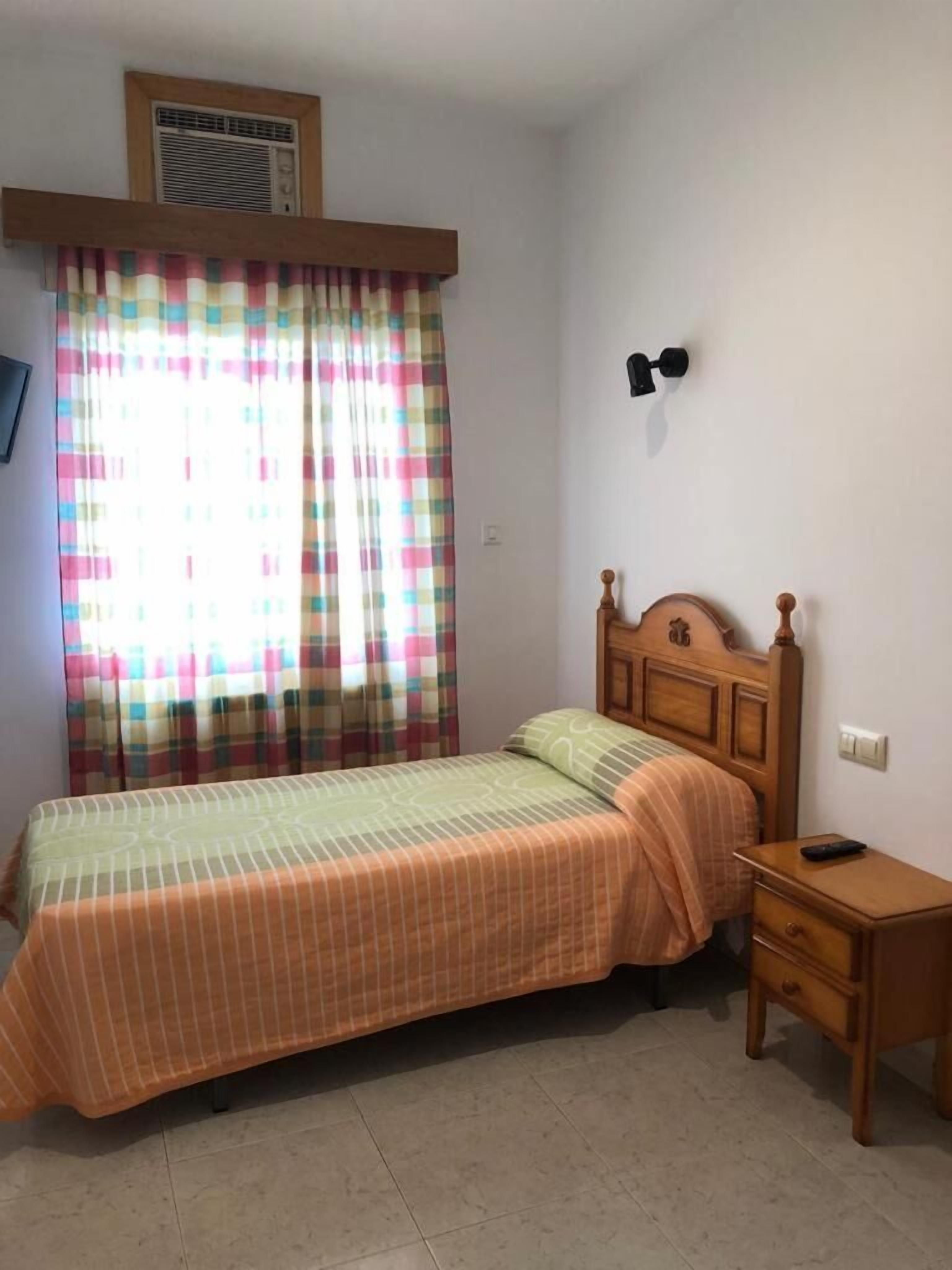 A bedroom with a bed, a nightstand, a wall-mounted lamp, and a window with curtains.