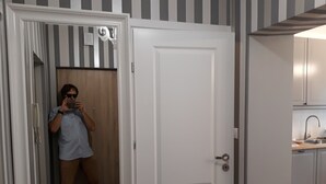 A person taking a selfie in a room with a striped wall, a wooden door, and a kitchen area visible through an open door.
