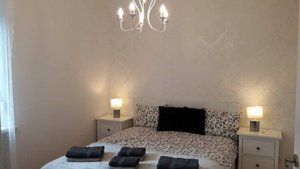 A bedroom with a chandelier, two bedside lamps, a patterned bedspread, a black pillow, and four folded towels.