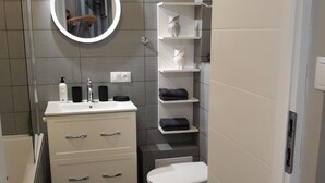A compact bathroom with a white vanity, a round mirror, a toilet, and a shelf with decorative items.