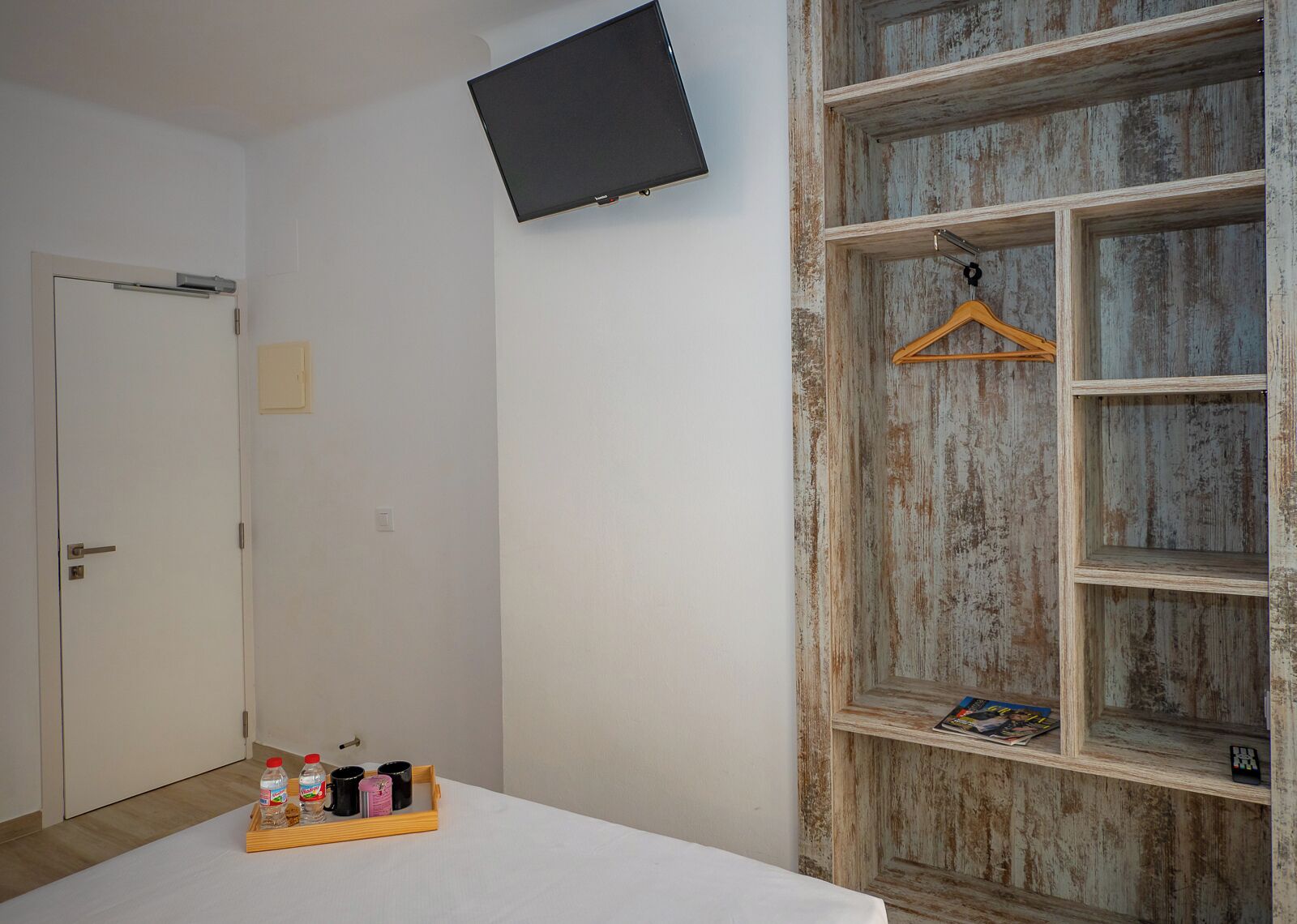 Une pièce avec une porte blanche, un téléviseur à écran plat fixé au mur, une étagère en bois avec un cintre, et un plateau avec des bouteilles et des tasses posé sur le sol.