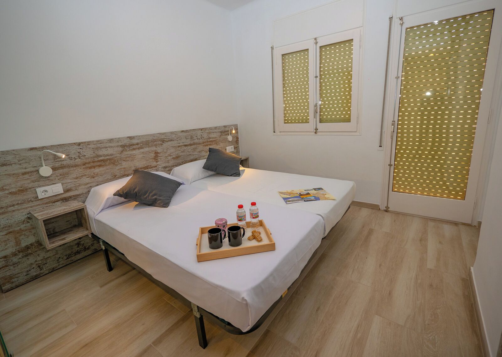 A neatly made bed with pillows, a bedside table with a tray of breakfast items, a wooden headboard, and a window with blinds.