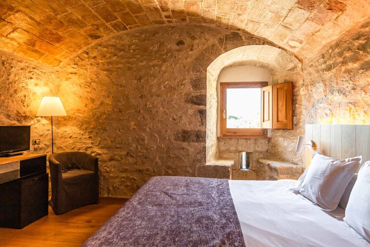 A room with a stone wall, a bed with white linens, a wooden desk with a lamp, and a television.