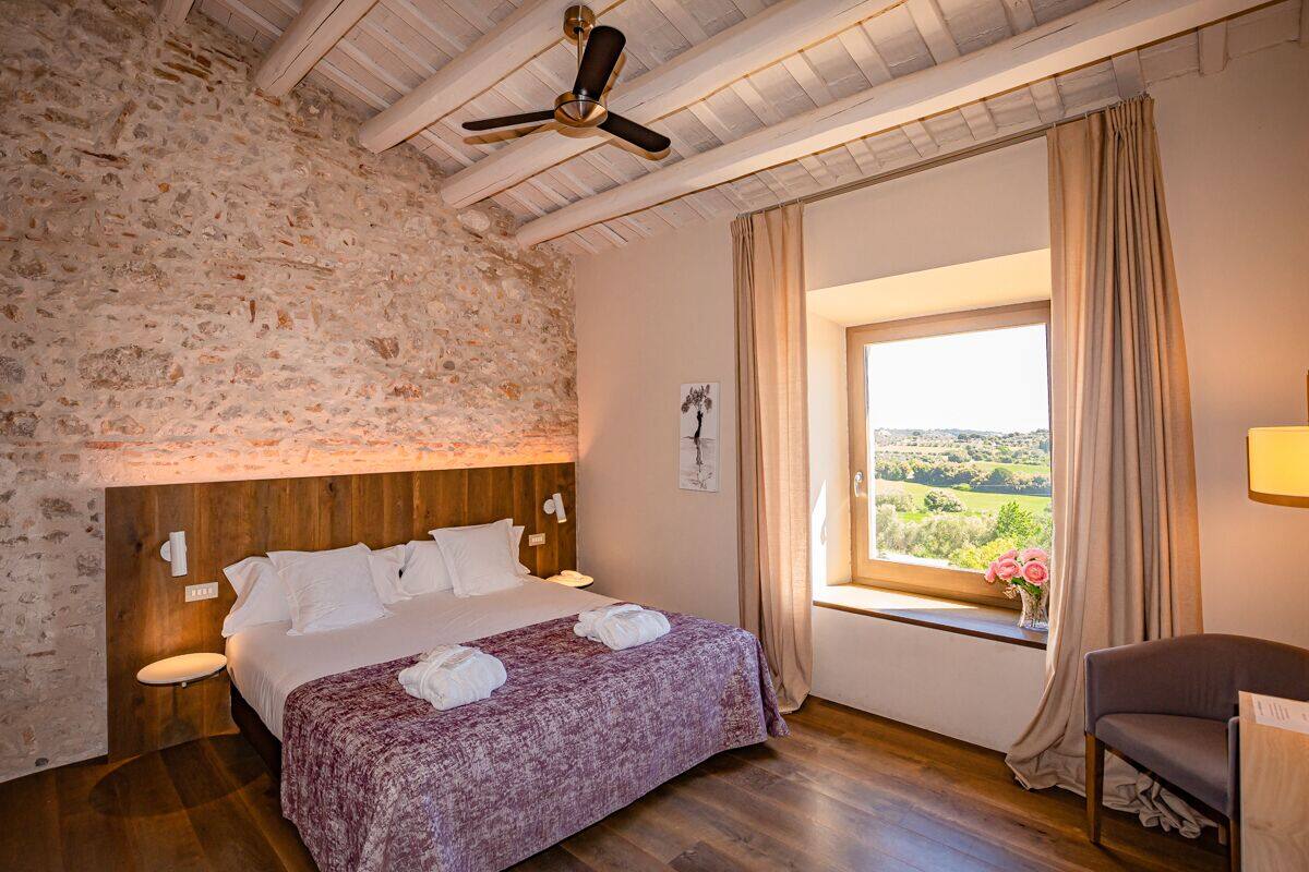 A bedroom with a stone wall, wooden ceiling beams, a bed with a purple bedspread, a window with a view, and a chair.