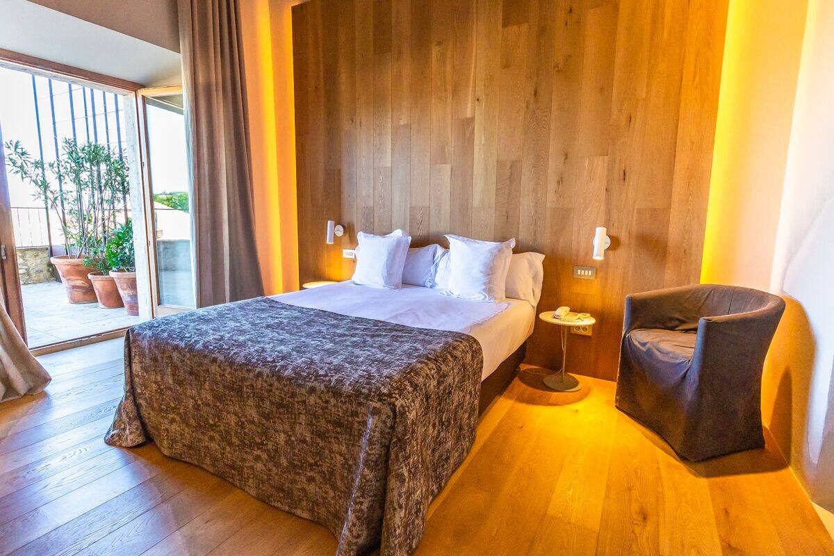 A bedroom with a bed, a chair, a nightstand, and a view of the outside.