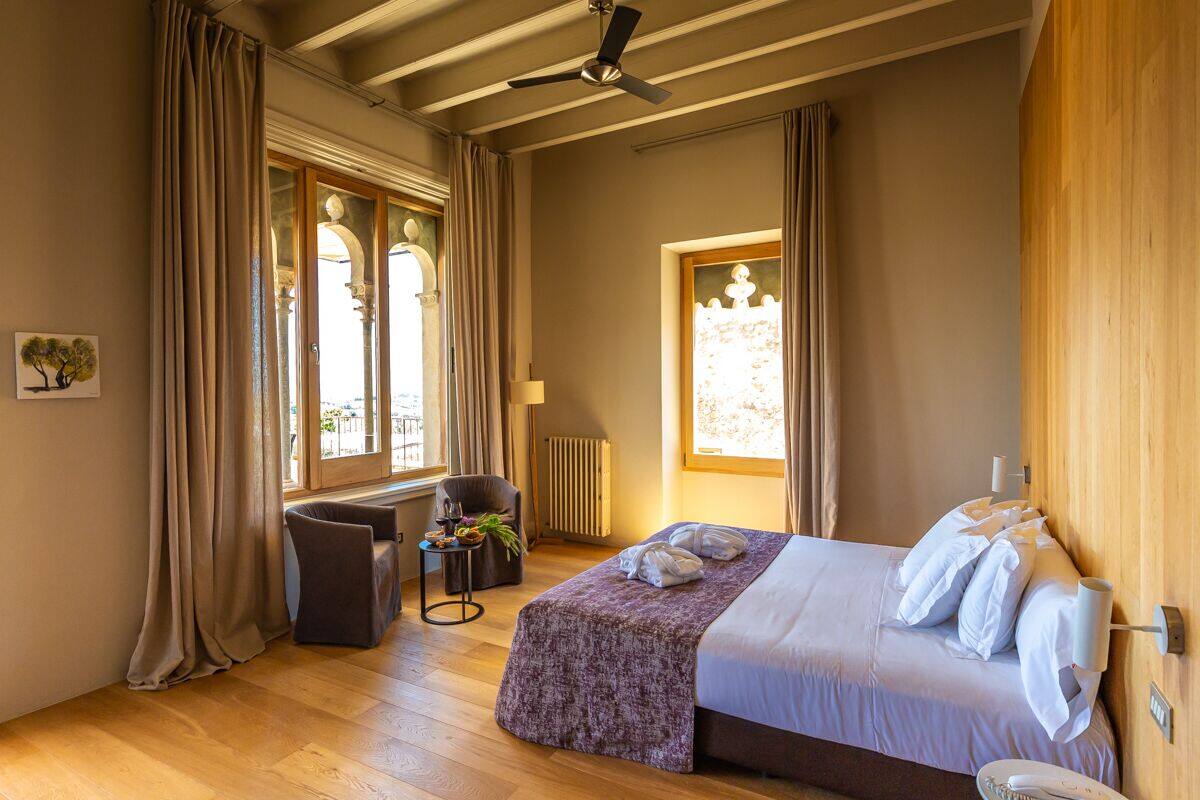 A hotel room with a bed, a chair, a small table with a plant, and a window with curtains.
