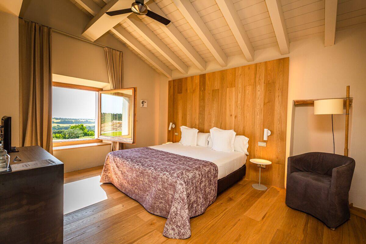 A bedroom with a large bed, wooden panel walls, a view through a window, a desk, and a chair.