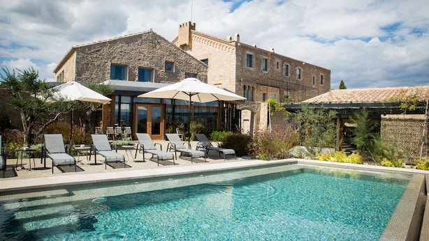 A poolside area with lounge chairs and umbrellas, a stone building with windows, and a clear blue pool. - Hotel Mas Bosch 1526 (Avinyonet de Puigventos)