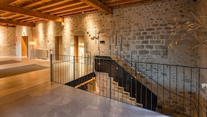 A spacious interior with a stone wall, a wooden ceiling, and a staircase with metal railings. - Hotel Mas Bosch 1526 (Avinyonet de Puigventos)