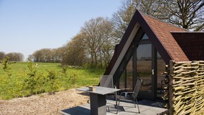 A modern, triangular-shaped building with a glass facade, a patio area with a table and chairs, and a view of a green field and trees in the background. - Restinn Medemblik (Wieringerwerf)