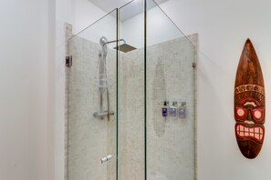Une douche fermée par des parois de verre, dotée d’un pommeau de douche et d’un panneau de commande, un masque décoratif en bois accroché au mur et un mur de douche recouvert de carreaux.