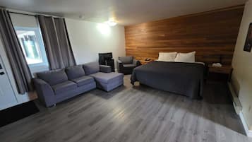 A modern hotel room with a grey sofa, a bed, a wooden accent wall, and a window with curtains.