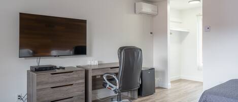 A modern hotel room with a wooden desk, a flat-screen TV, a grey office chair, a mini-fridge, and a bed with grey bedding.