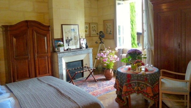 A bedroom with a bed, a wooden wardrobe, a fireplace, a table with a vase of flowers, and a window with curtains. - Domaine Mongiron (Nerigean)