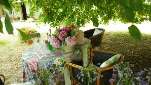 An outdoor dining area with a table set for two under a tree, surrounded by wicker chairs with cushions. - Domaine Mongiron (Nerigean)