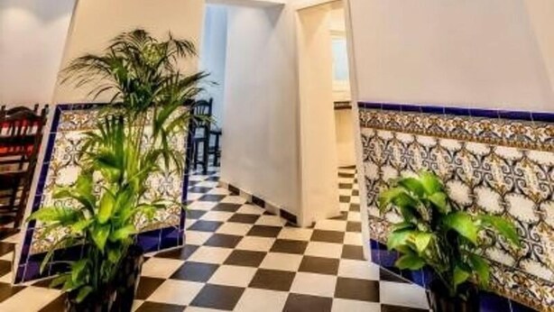 A high-ceilinged interior with a checkered floor, a staircase leading to an upper level, and a decorative wall with a patterned tile border. - Hostel A2C - Adults Only (Seville)